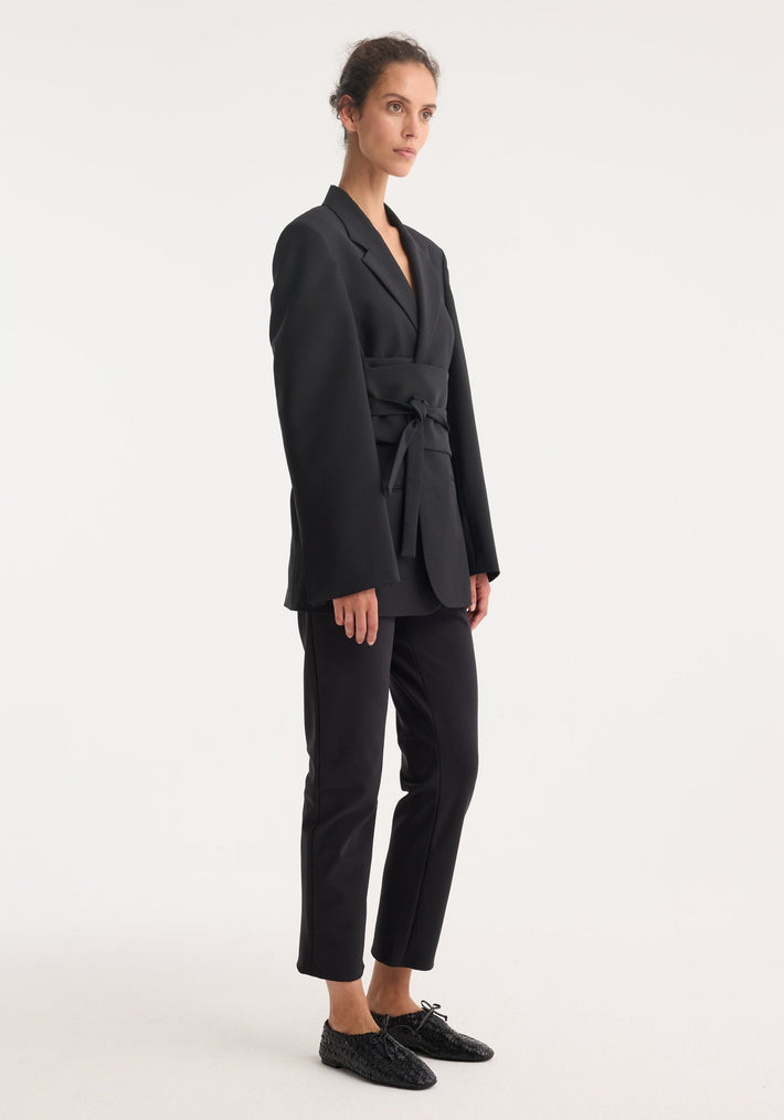 Woman models a Kimono belted blazer in black, featuring a wrap sash belt and tailored fit, paired with slim trousers and lace-up loafers.