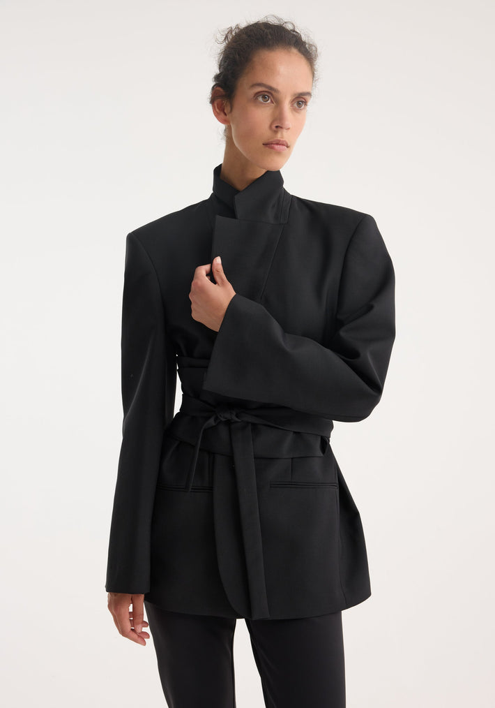Kimono belted blazer in black, featuring an asymmetrical high-collar and wrap sash belt, worn by a slender woman with hair casually pulled back.