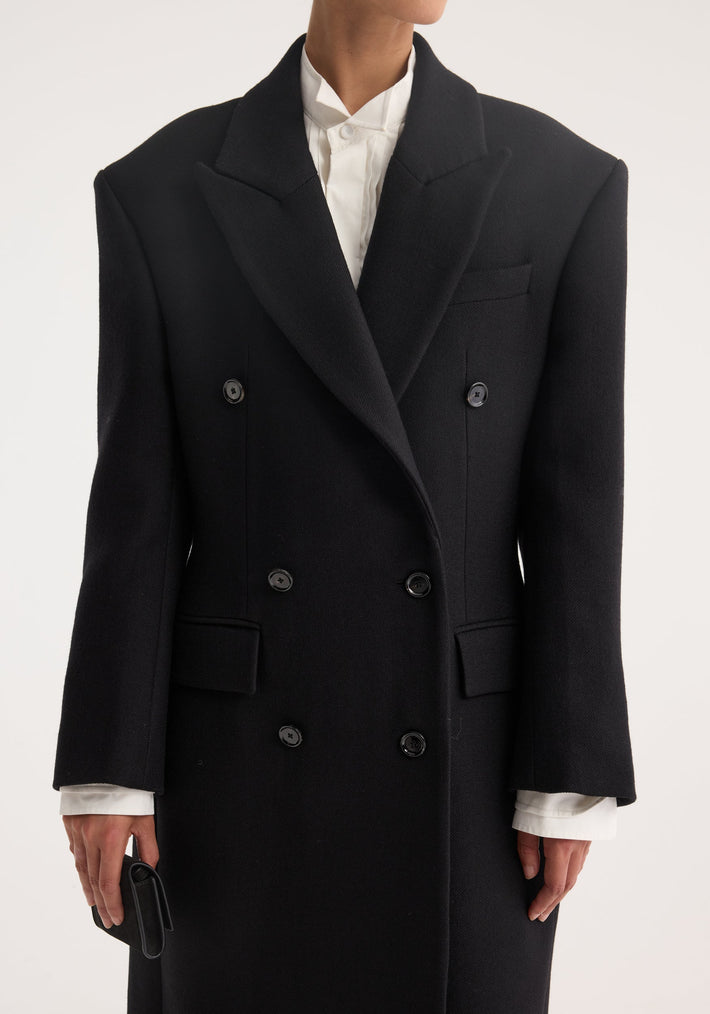 Hourglass wool coat in black, double-breasted with six buttons, worn over a white shirt. The model holds a small black wallet in hand.