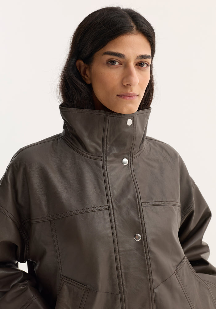 Person wearing Reinvented leather jacket | earth, featuring high snap-button collar, visible stitching, and patch pockets. Relaxed bomber silhouette with soft leather and oversized fit.