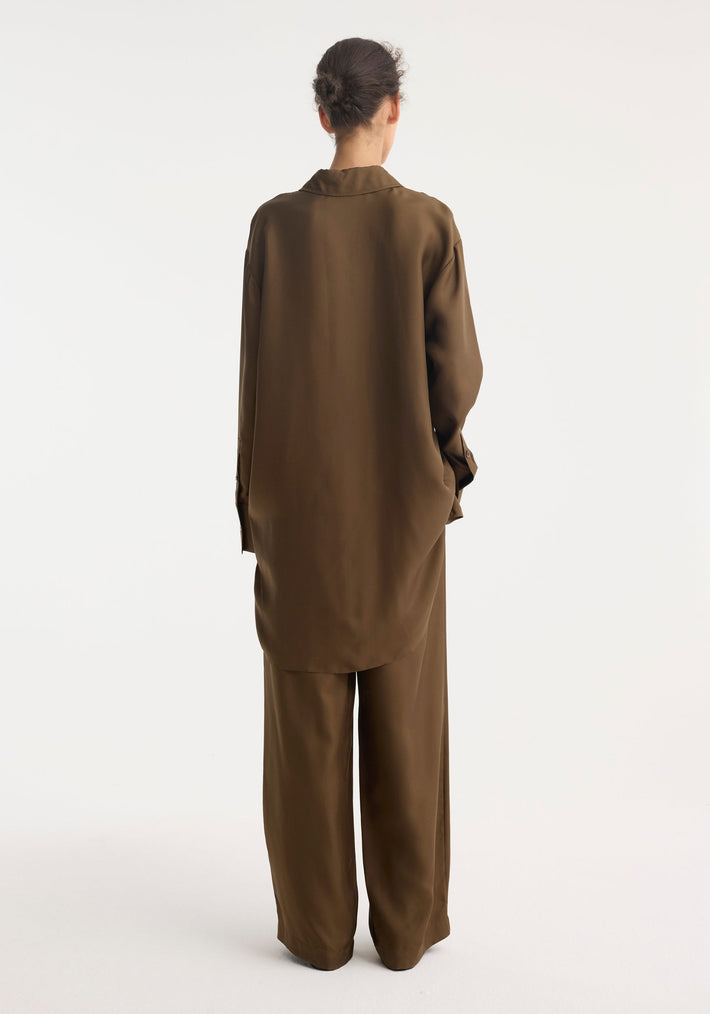 Person wearing the Signature elongated silk shirt in forest, showcasing its oversized fit and statement collar from behind, paired with matching wide-leg trousers.