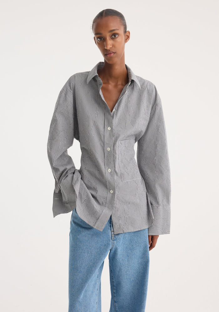 Model wearing a Crushed checked shaped shirt with a mini grey check pattern, featuring a waisted silhouette and exaggerated cuffs, paired with loose-fit denim jeans.