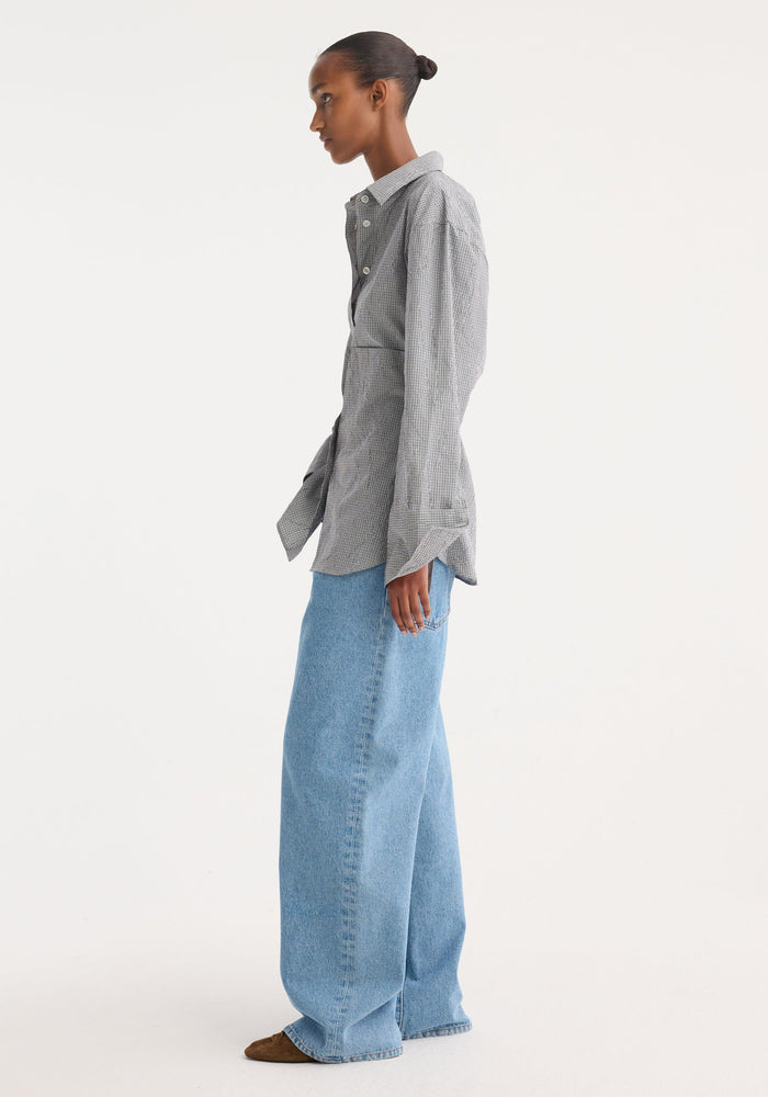 Woman in a Crushed checked shaped shirt with asymmetrical tie detail, paired with wide-leg jeans and brown suede shoes, exuding a minimalist style.