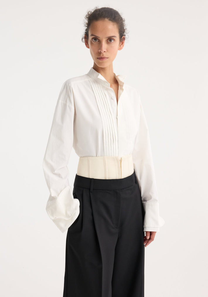 Model wearing Tuxedo wired shaped shirt in optic white, featuring pintucked front, stand collar, statement cuffs with a cream corset belt and black wide-leg trousers.