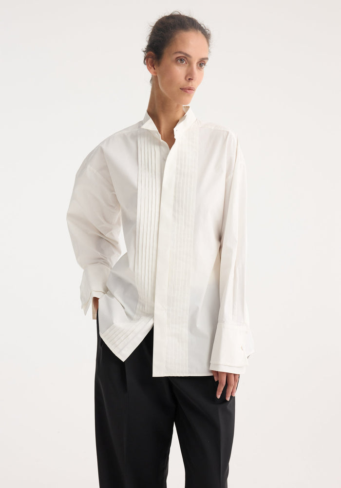 Model in a Tuxedo wired shaped shirt | optic white featuring subtle pleats, stand collar, statement cuffs, and curved hem, paired with black wide-leg trousers.