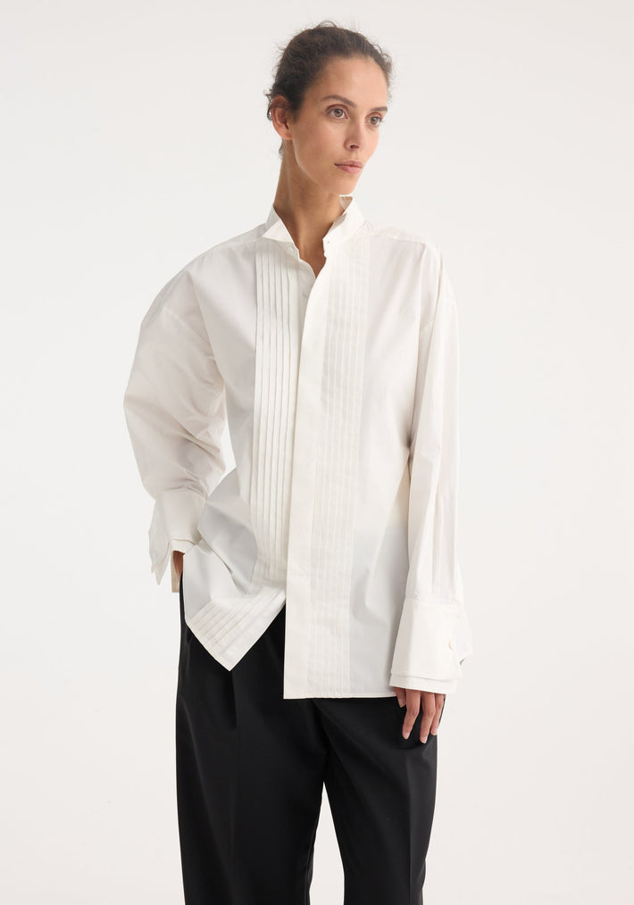 Model in a Tuxedo wired shaped shirt | optic white featuring subtle pleats, stand collar, statement cuffs, and curved hem, paired with black wide-leg trousers.