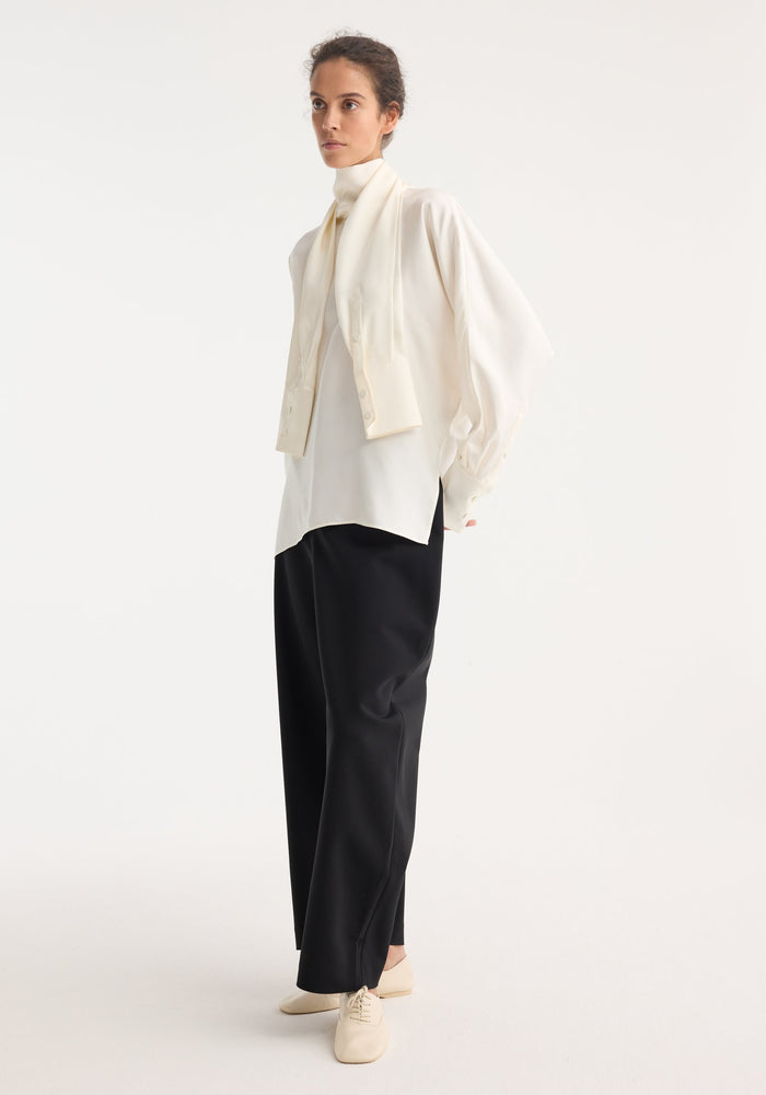 Model wearing Foulard silk top with draped sleeves, layered over a high-neck top, paired with loose black trousers and cream lace-up flats.