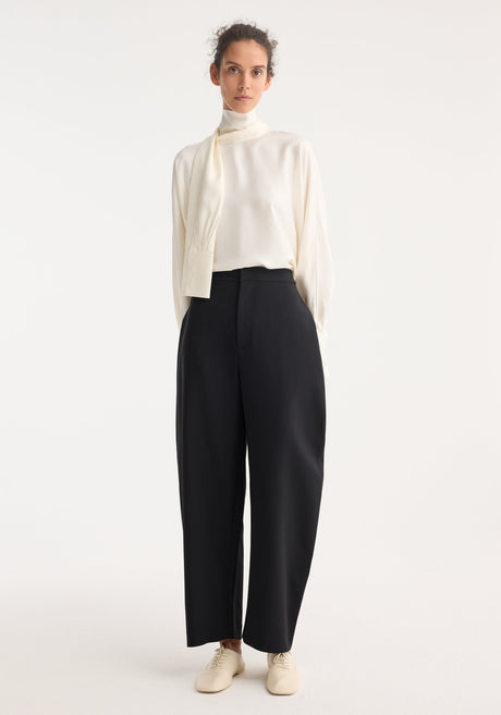 Woman models Foulard silk top, featuring draped sleeves and scarf tie, paired with high-waisted black trousers and lace-up flats, showcasing a minimalist, tailored look.