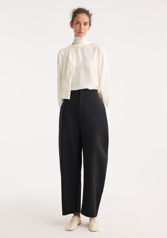 Woman models Foulard silk top, featuring draped sleeves and scarf tie, paired with high-waisted black trousers and lace-up flats, showcasing a minimalist, tailored look.