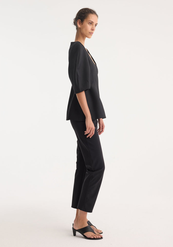 Young woman in a Sculptural top | black, featuring a deep scoop neckline and elbow-length balloon sleeves, paired with straight-leg pants and heeled slide sandals.
