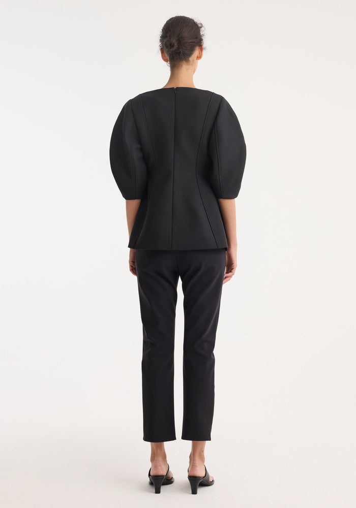 Woman in rear-view wearing Sculptural top | black with puff sleeves, peplum waist, paired with slim-fit trousers and open-toe sandals, highlighting the top's structured silhouette.