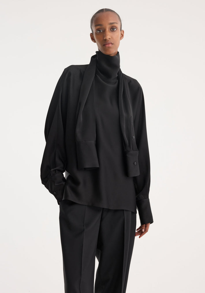 Model wearing Foulard satin top with draped sleeves, extended turtleneck as a scarf, paired with tailored trousers, showcasing a minimalist, modern silhouette.