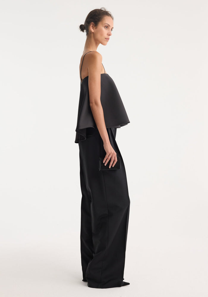 Tall woman in profile wearing a black peplum satin top with adjustable spaghetti straps, wide-leg trousers, and holding a small rectangular clutch.