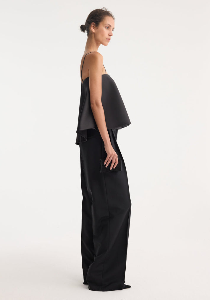 Tall woman in profile wearing a black peplum satin top with adjustable spaghetti straps, wide-leg trousers, and holding a small rectangular clutch.