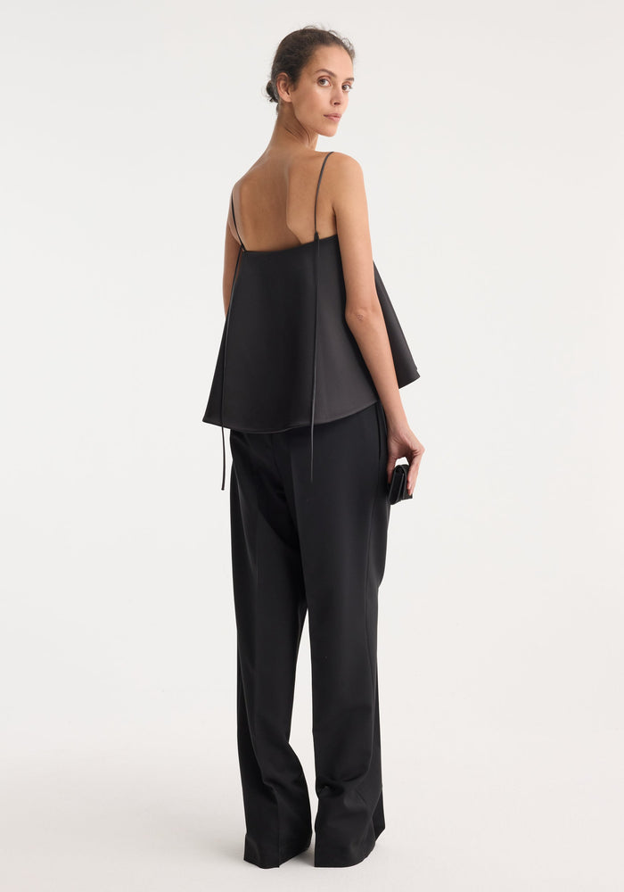 Woman in profile wearing the Peplum satin top with thin straps, flaring peplum, holding a small clutch, showcasing the elegant silhouette.