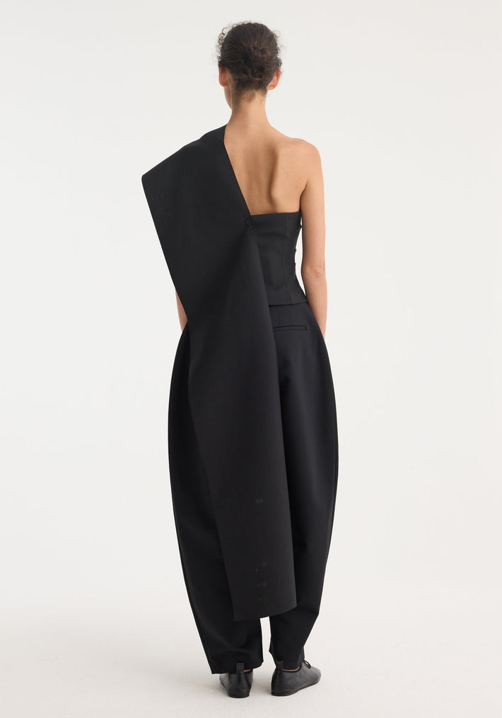 Model in a One-shoulder cape top | black, featuring a strapless bustier, asymmetric draped sash, paired with voluminous high-waisted trousers and lace-up shoes.