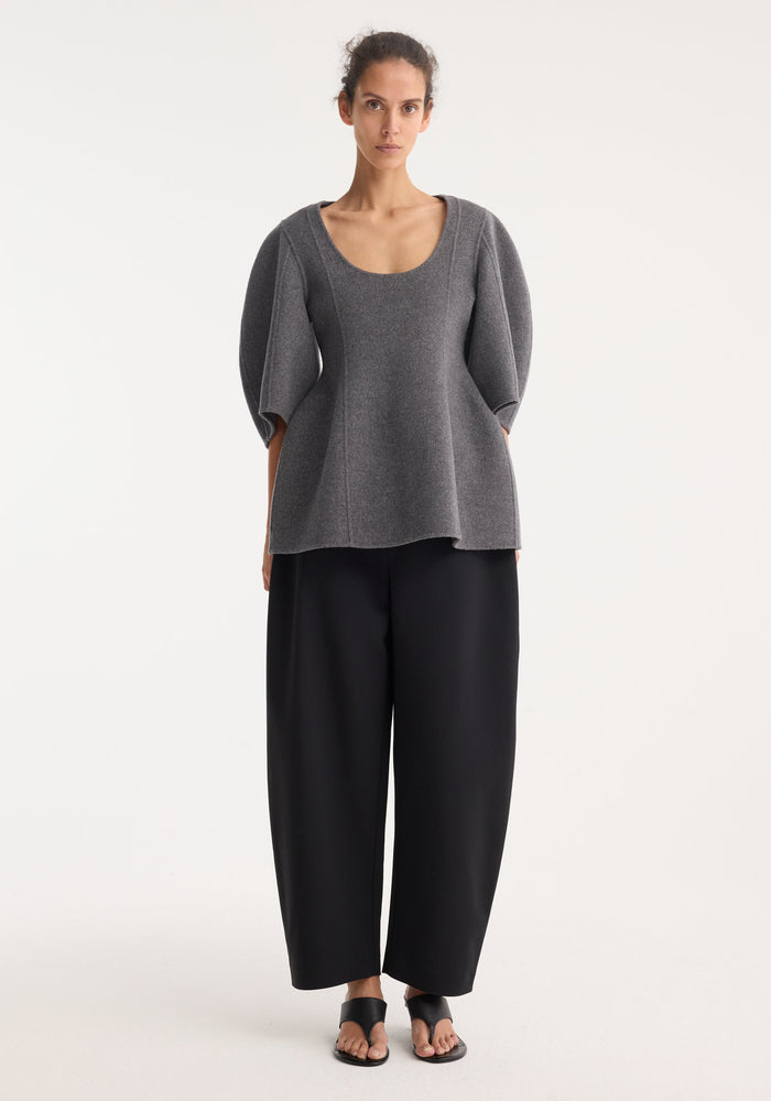 Model wearing Sculptural double-faced top in mid grey melange with a scoop neckline, voluminous elbow-length sleeves, and peplum flare, paired with black wide-leg trousers and sandals.