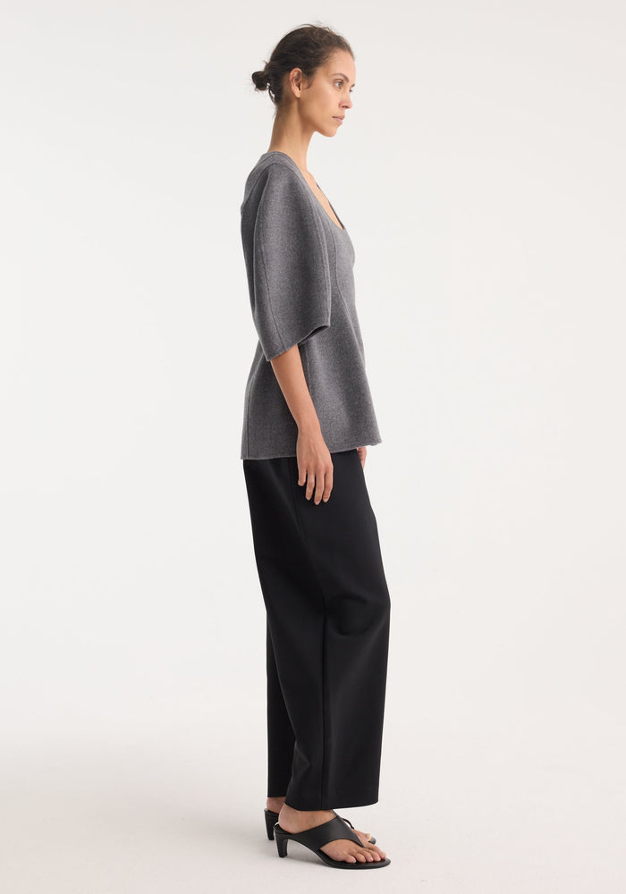 Model in a mid grey melange sculptural double-faced top, featuring a deep scoop neck, elbow-length balloon sleeves, and a structured peplum silhouette, paired with black trousers.