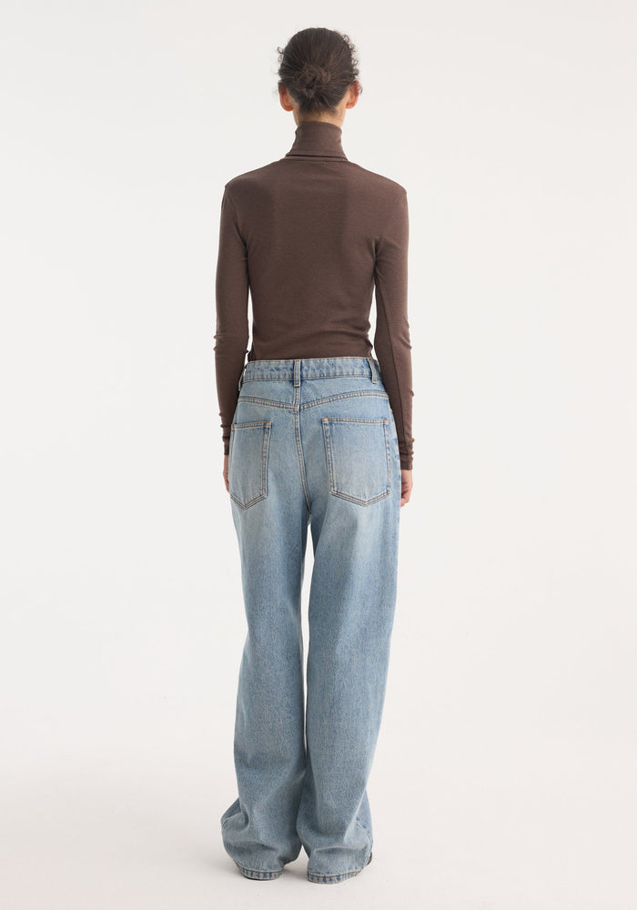 Person wearing Fine merino turtleneck in brown melange, fitted with long sleeves, paired with high-waisted, wide-leg light-wash jeans, hair in a bun.