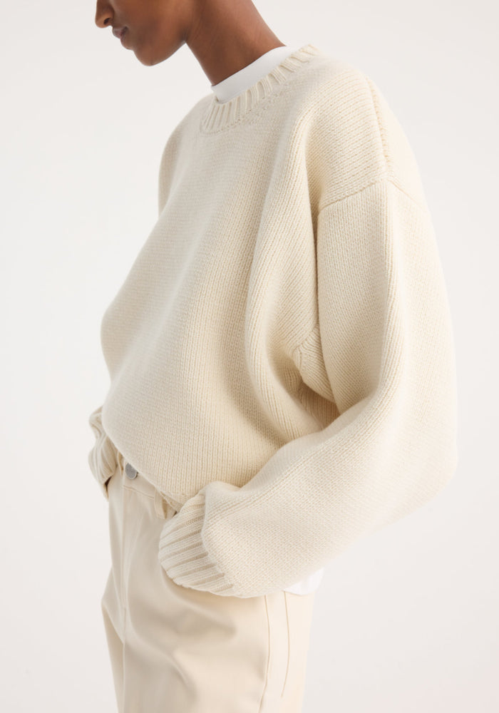 Person wearing the Sculptural compact crewneck in off white, a loose rib-knit sweater with dropped shoulders, tucked into high-waisted trousers, hands in pockets.