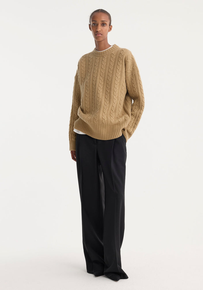 Model in a camel Cable Knit Jumper, oversized fit, round neckline, paired with wide-leg black trousers; showcasing minimalist, modern style.