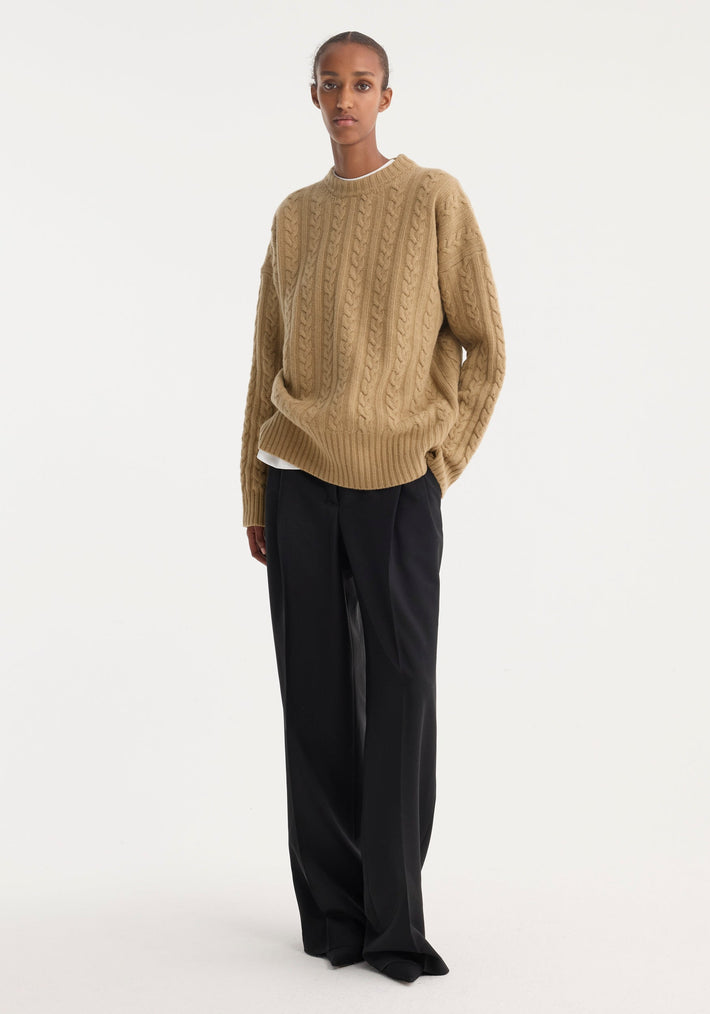 Model in a camel Cable Knit Jumper, oversized fit, round neckline, paired with wide-leg black trousers; showcasing minimalist, modern style.