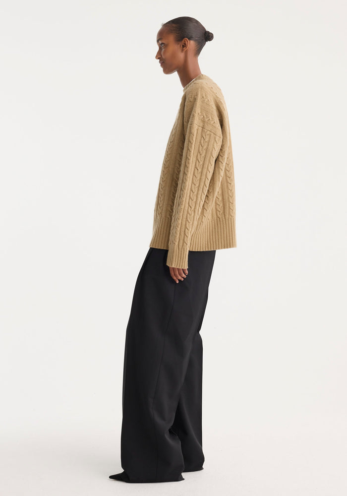 Person in oversized Cable knit jumper | camel, with dropped shoulders and round neckline, paired with wide-leg trousers. Hair in bun, showcasing modern fashion style.