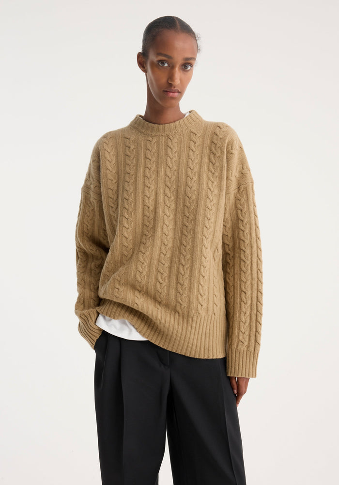 Young woman wearing an oversized Cable knit jumper | camel with drop shoulders and ribbed details, paired with black pleated trousers, standing in a relaxed pose.