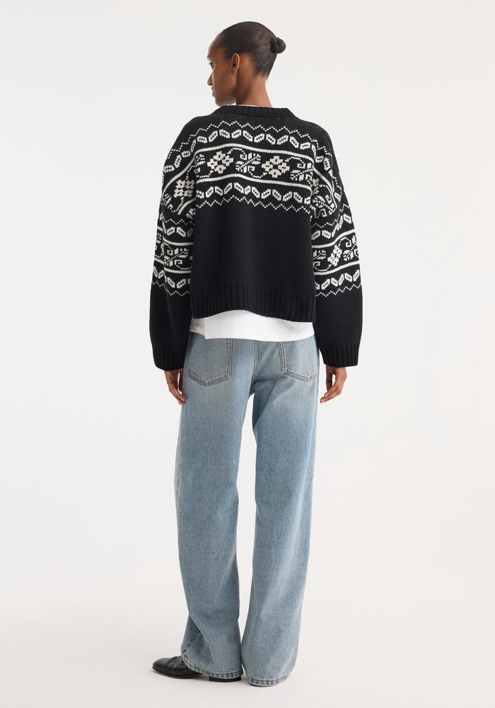 Woman wearing Fair-isle knitted jumper | black, featuring a classic pattern, boxy fit, slightly cropped. Paired with light-wash wide-leg jeans and slip-on shoes.