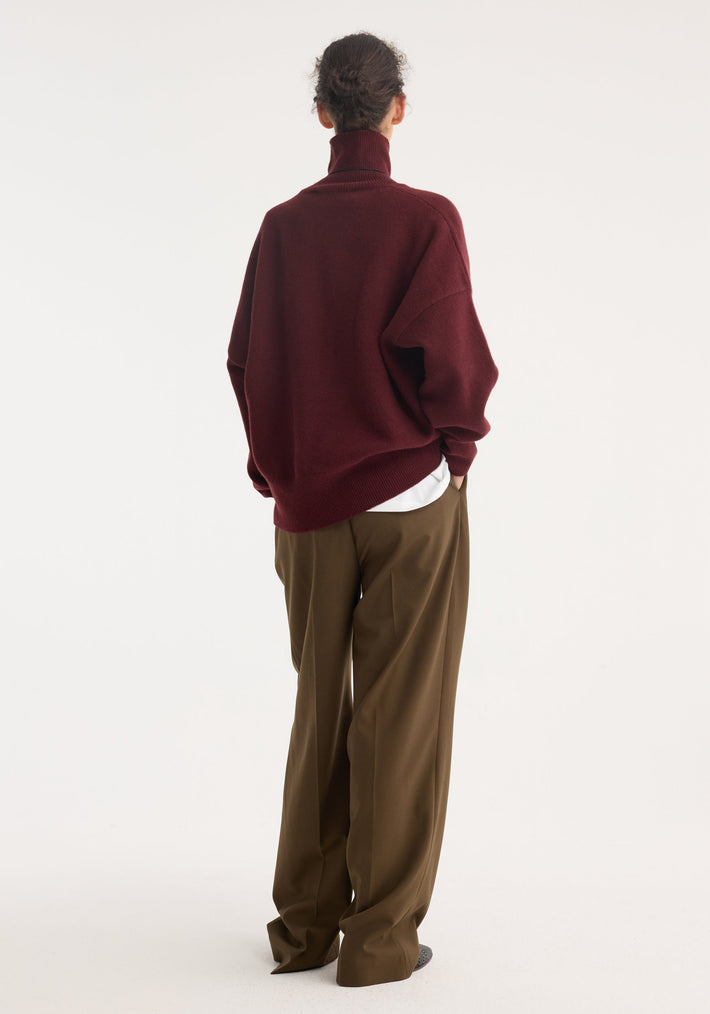 Classic turtleneck jumper in burgundy, oversized with drop shoulder, layered over a white shirt, paired with olive-brown wide-leg trousers, creating a relaxed silhouette.