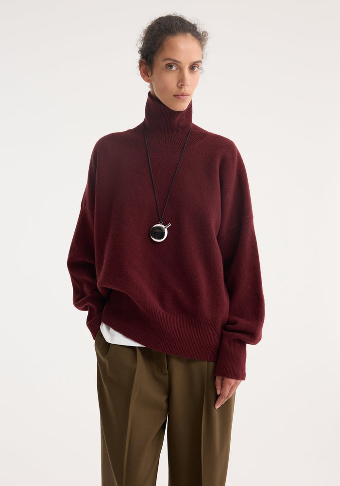 Model wearing a Classic turtleneck jumper | burgundy, loose-fit, over a white undershirt, with olive-brown wide-leg trousers, accessorized with a dark cord necklace and pendant.