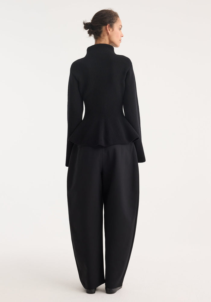 Woman wearing Sculptural knitted top with high collar, elongated sleeves, and peplum flare, paired with wide-leg trousers, viewed from behind.
