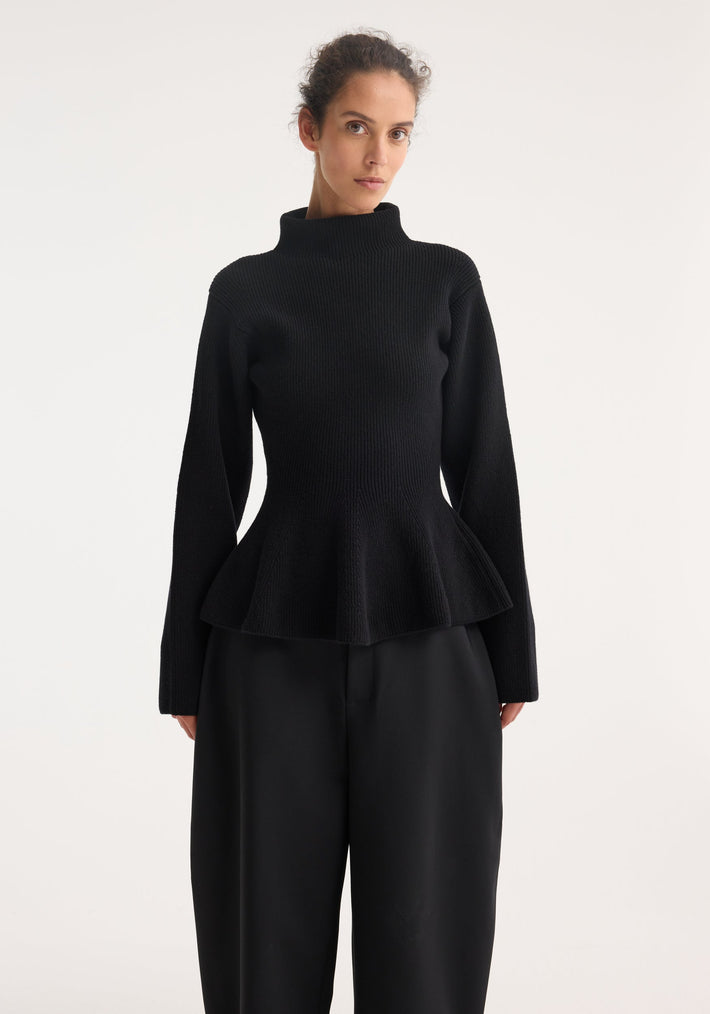 Woman in a Sculptural knitted top with high turtleneck, long sleeves, and peplum waist, paired with wide-leg trousers, showcasing elegant knitwear craftsmanship.