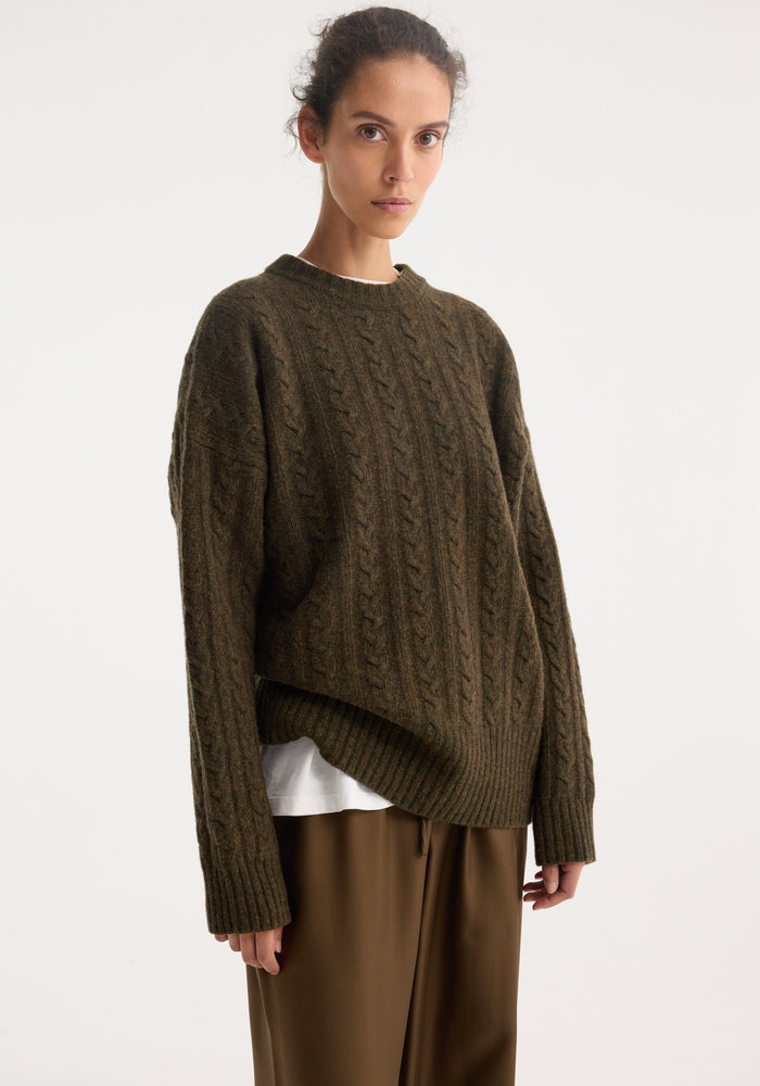 Woman wearing Cable knit jumper | forest melange, oversized wool pullover with cable texture, crew neckline, and dropped shoulders, layered over a white T-shirt.