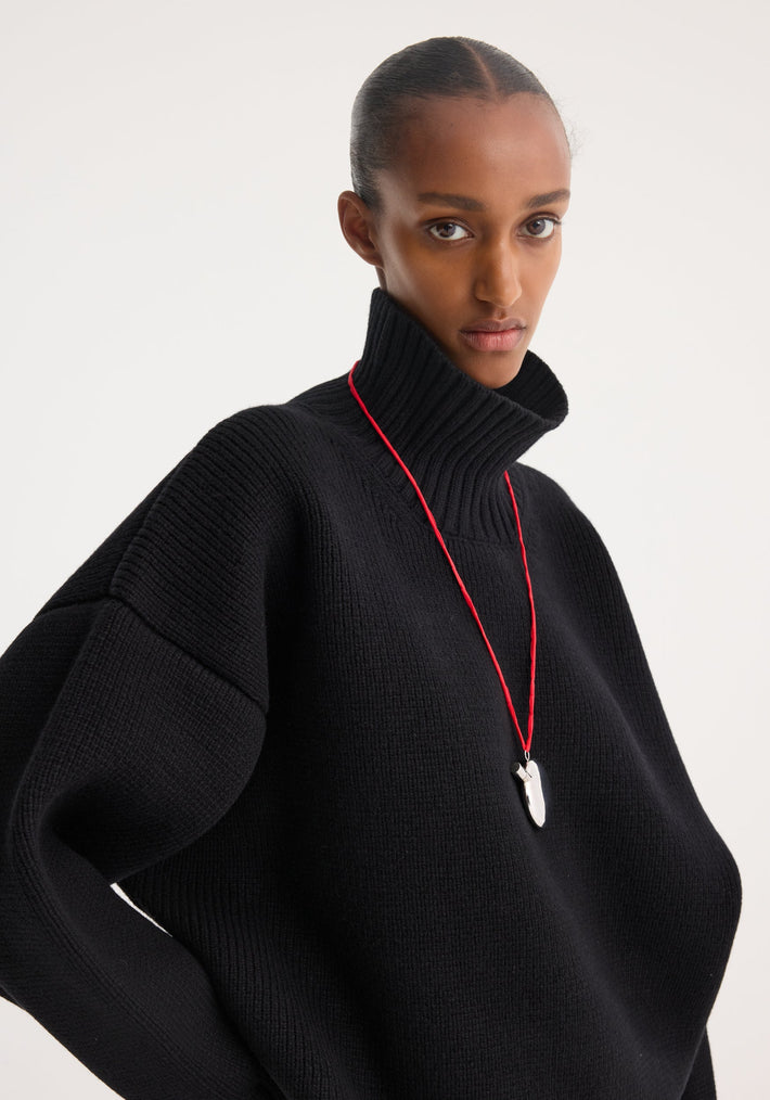 Sculptural compact turtleneck in black, worn by a slender model. Features chunky knit, relaxed fit, drop shoulder. Silver pendant accessory visible.