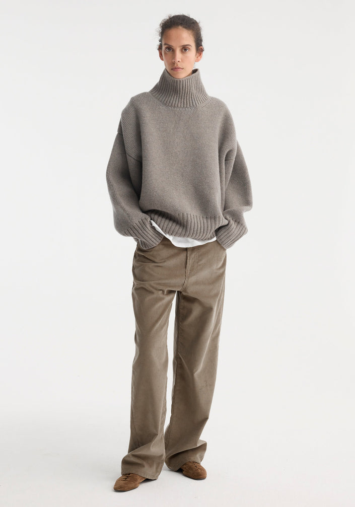 Woman wearing Sculptural compact turtleneck | clay melange, an oversized grey knit with drop shoulders, over loose taupe trousers, styled with brown suede lace-up shoes.