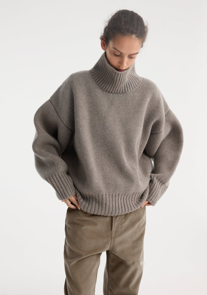 Person wearing Sculptural compact turtleneck | clay melange, showcasing its relaxed fit and drop shoulder design, paired with loose corduroy trousers for a cozy look.