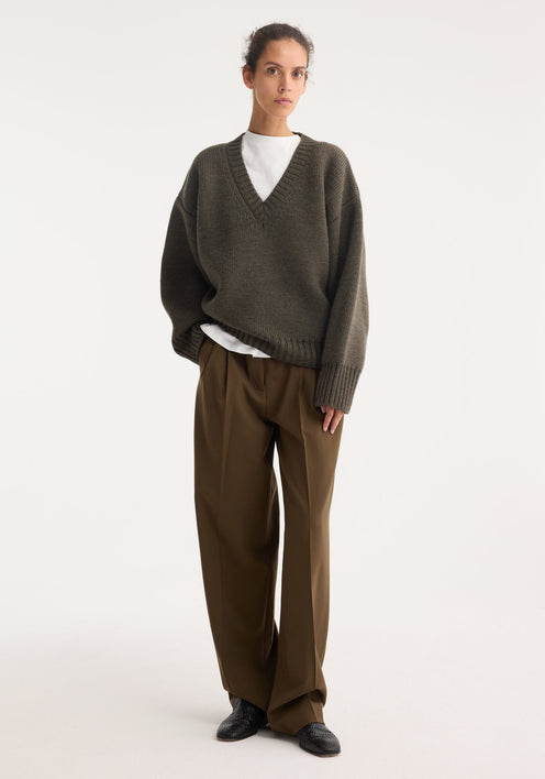 Model wearing Sculptural compact v-neck | forest melange, an oversized olive-green V-neck sweater, over a white T-shirt, paired with brown wide-leg trousers and black loafers.