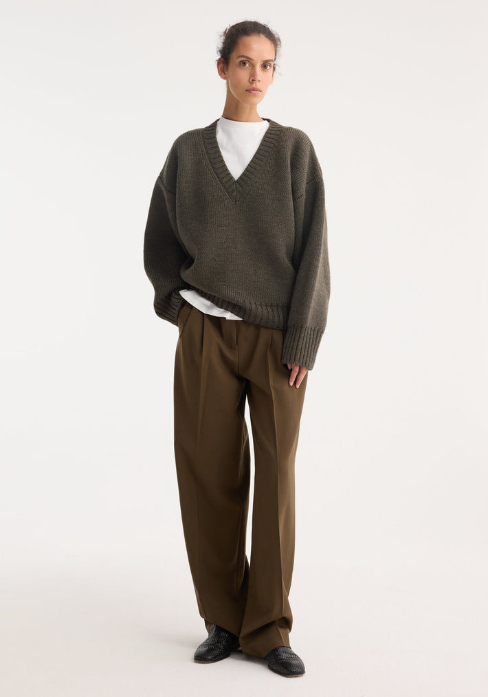Model wearing Sculptural compact v-neck | forest melange, an oversized olive-green V-neck sweater, over a white T-shirt, paired with brown wide-leg trousers and black loafers.