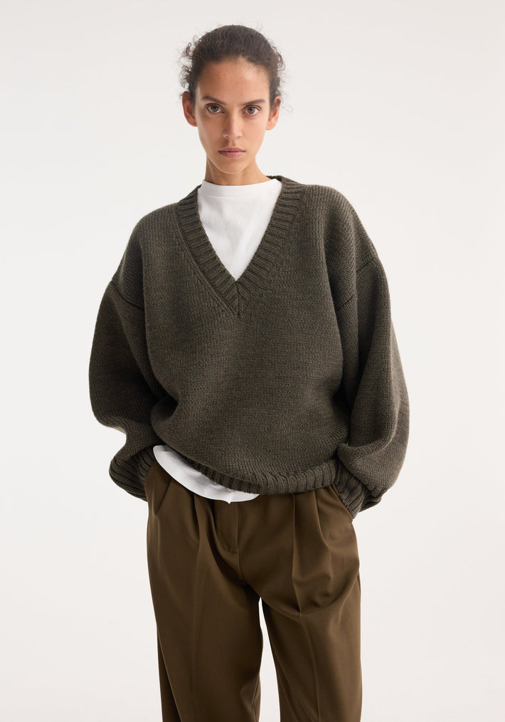 Person wearing Sculptural compact v-neck | forest melange, featuring an oversized V-neck knit sweater over a white T-shirt with loose-fit brown trousers, hands in pockets.