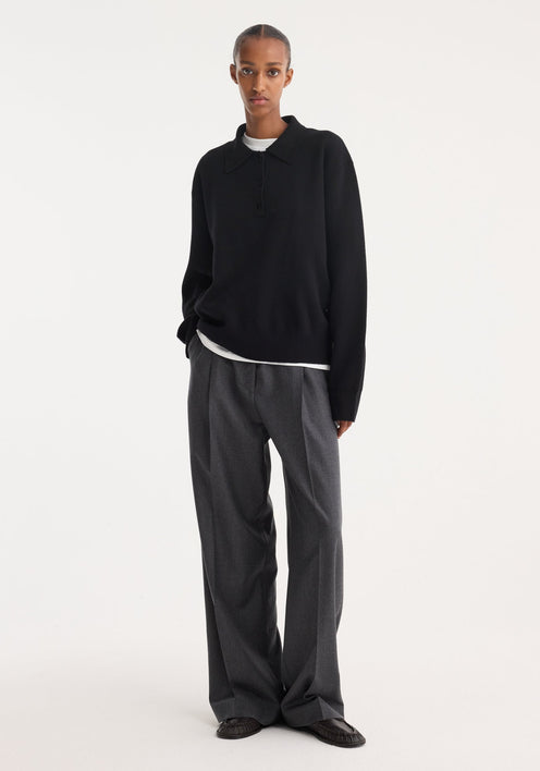 Tall person wearing a black Compact knitted polo with a three-button placket, over a white T-shirt, paired with wide-leg trousers and loafers.