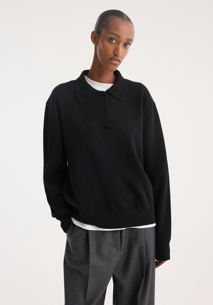 Tall person wearing a black Compact knitted polo with a three-button placket, over a white T-shirt, paired with wide-leg trousers and loafers.