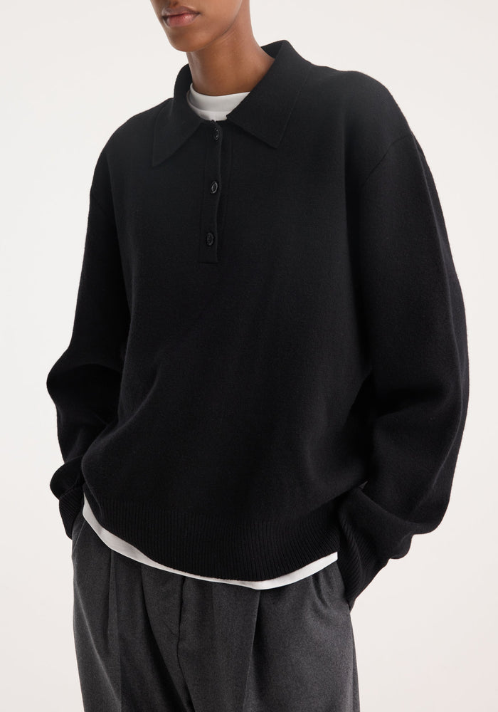 The model wears a Compact knitted polo in black, featuring a relaxed fit, long sleeves, and a three-button placket, layered over a white T-shirt.