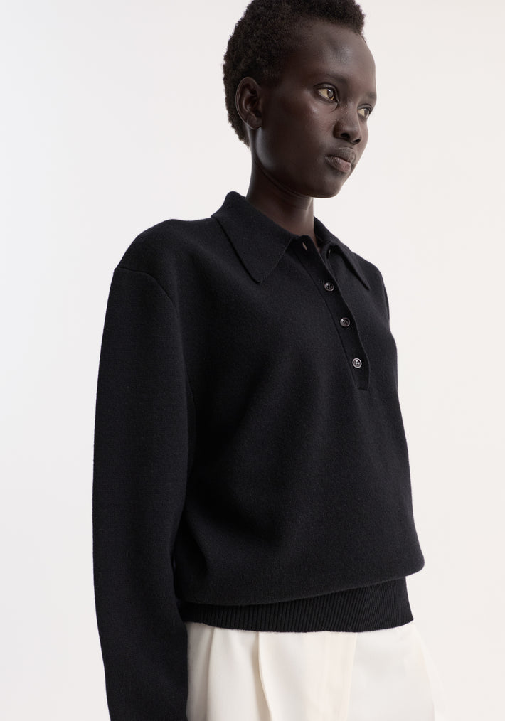Person in a Compact knitted polo | black with a 3-button front, rounded drop shoulder, and structured collar, paired with loose white trousers.