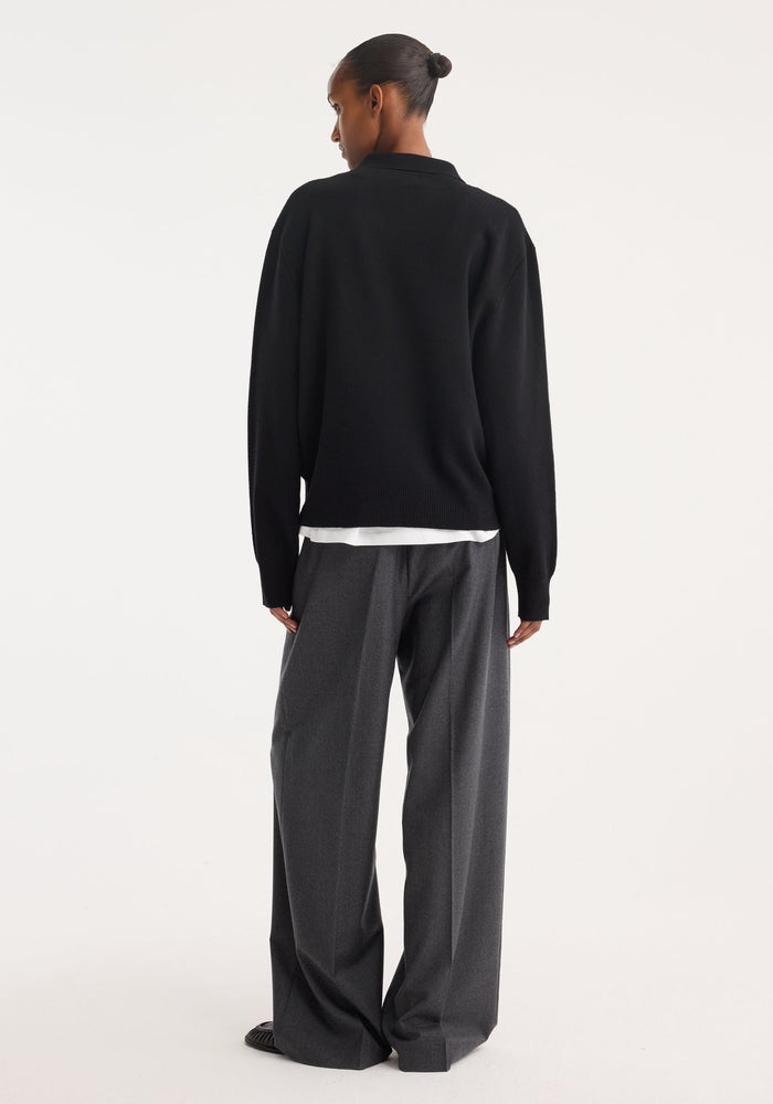 Person wearing a black Compact knitted polo with a collar and 3-button front, layered over a white shirt, paired with gray high-waisted trousers and black loafers.