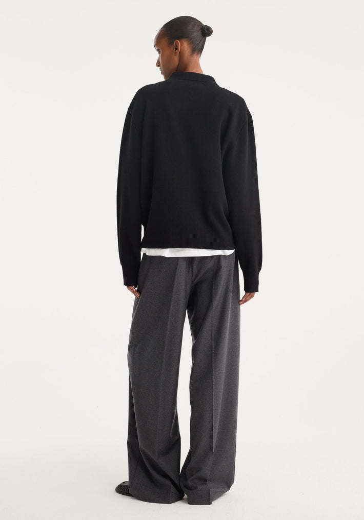 Person wearing a black Compact knitted polo with a collar and 3-button front, layered over a white shirt, paired with gray high-waisted trousers and black loafers.