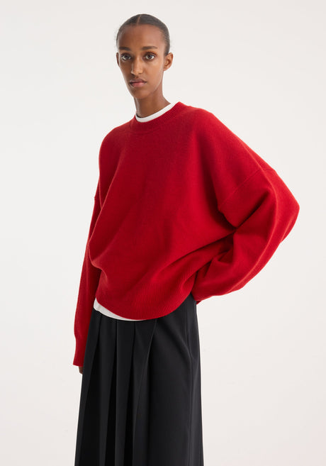 Person wearing a Boiled wool crewneck | rosso, featuring a relaxed fit and drop shoulders, paired with a black pleated midi skirt and white kitten-heel mules.