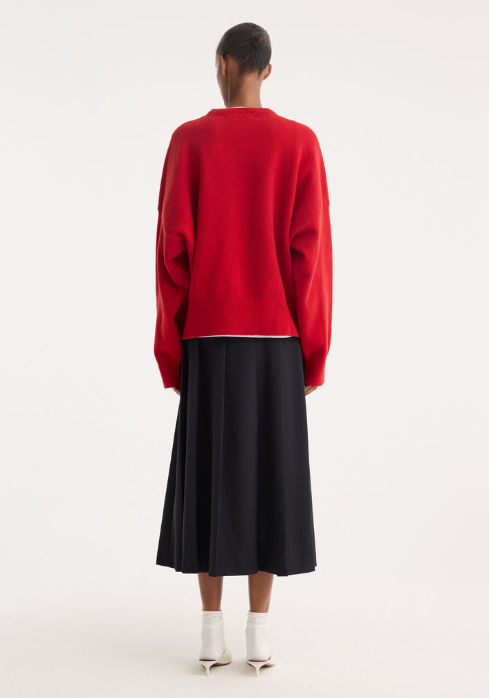 Person wearing Boiled wool crewneck | rosso, a loose-fitting red sweater, with a high-waisted black pleated skirt, white ankle socks, and pointed white heels.