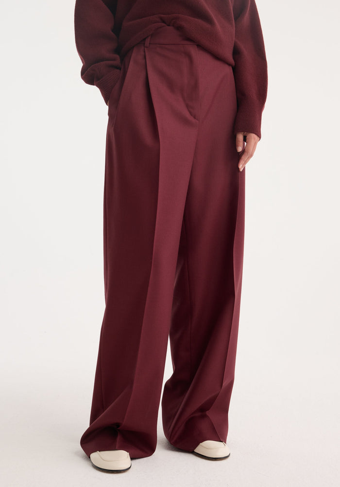 Mid-waist tailored trousers in burgundy, featuring front pleats, side pockets, and a wide-leg design, crafted from soft virgin wool.