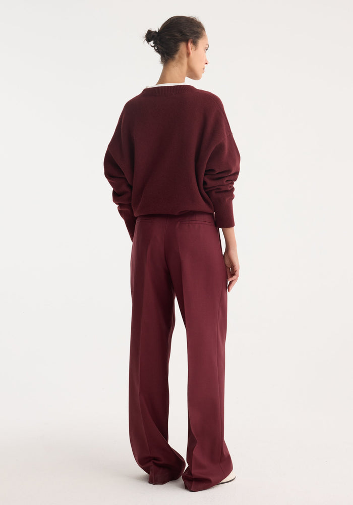 Person wearing Mid-waist tailored trousers in burgundy, featuring a wide-leg design with double pleats, seen from behind with matching sweater and white shoes visible.